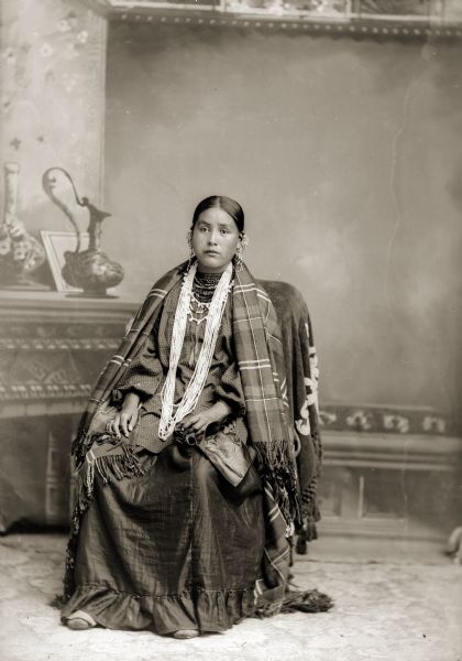 Full-length studio portrait of a Ho-Chunk woman, Lucy Decorah (AhHoRaPaNeeWinKah), posing sitting in front of a painted backdrop. She is wearing a wool shawl over a dress, white bead necklaces, and many silver ear bobs.