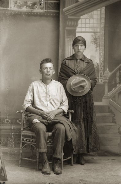 Full-length studio portrait of a Ho-Chunk man and woman in front of a painted backdrop. The man is posing sitting, and the woman is standing beside him and is wearing a scarf on her head and a shawl around her shoulders. She is holding the man's hat in front of her. They are both wearing modern dress. Identified as John Dick (DoWjhGeNaNoKah), and his wife Mary Manly-Mallory (Breeze) Dick (HaJaKeePaNaWinKah). They had a son named Melvin G. Dick who was born in 1923. 

According to Tomah Indian School's Indian Census Roll in 1933, the Dicks were under the jurisdiction of Winnebago, Nebraska. They were possibly members of the Wisconsin Rapids band, and their surname was formerly Breeze according to the notes of Jackson County Historical Society, Wisconsin.