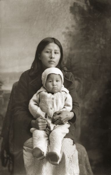 Three-quarter length studio portrait of a Ho-Chunk woman posin sitting and holding a small child in winter clothes in front of a painted backdrop. Identified as Bertha Greengrass Blackdeer (HaukSeGoHoNoKah), the wife of Irwin Blackdeer, with her niece, Bernice, the daughter of Ruth Greengrass Cloud.