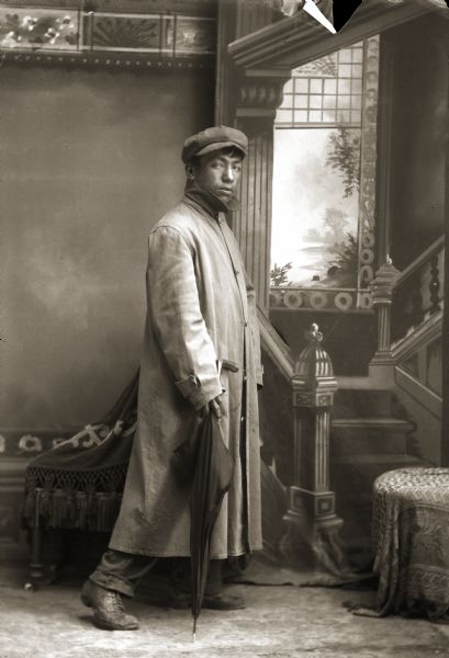 Full-length studio portrait of a Ho-Chunk man, Will (Willy) Goodvillage (MaHayEKeReNaKAh), posing standing in front of a painted backdrop near a chair. He is holding an umbrella and is wearing a long overcoat and a hat. Not many of Van Schaick's Ho-Chunk photographs are this animated. Will was blind, and as an old man, his uncle, John Swallow (MonKeSaKaHepKah), who was a few years older, cared for him and brought him to feasts.