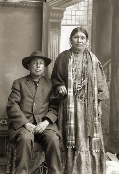 Three-quarter length studio portrait of a Ho-Chunk man and woman in front of a painted backdrop. The man is posing sitting and is wearing a suit and hat. The woman is standing on the right and is wearing beads, earrings, and a plaid shawl. Identified as Edward Funmaker (WaGeSeNaPeKah) and Mary Johnson (ENooKah).