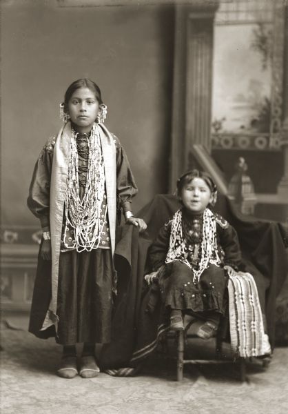 Full-length studio portrait of two young Ho-Chunk girls, one posing standing and the other sitting, both wearing bead necklaces and possessing shawls in front of a painted backdrop. They are dressed in their finest outfits. The girl on the right is seated next to a Ho-Chunk blanket with silk applique "beaver tail" design. The girl on the left is Mabel White Blackhawk St. Cyr.