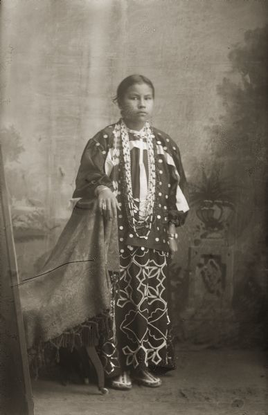 Full-length studio portrait in front of a painted backdrop of a Ho-Chunk woman, Mary Edith Wallace Vazquez, posing standing with her arm resting on a stuffed chair. She is wearing a Ho-Chunk woman's outfit in full regalia, including long necklaces, beaded shirt, and a very rare floral beaded skirt.