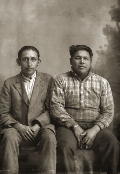 Three-quarter length studio portrait of two Ho-Chunk men posing sitting in front of a painted backdrop. The man on the left, John Payer, wearing the suit jacket may be a white man. The man on the right, Louis Johnson (HaNuKaw), is wearing a plaid shirt with an up-turned collar and a bandaged index finger on his left hand. John came from Nebraska, and Louis Johnson is listed on the Wisconsin tribal rolls.