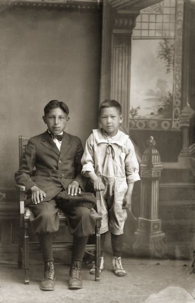 Studio portrait of two Ho-Chunk boys in front of a painted backdrop. The older boy, William Crow is posing sitting on the left wearing a suit, breeches, and a bow tie. The younger boy posing standing on the right, Frances Cassiman, is wearing a sailor suit.