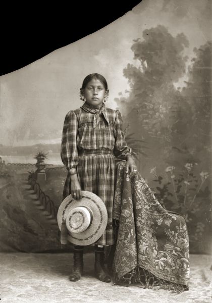 Studio portrait in front of a painted backdrop of a Ho-Chunk girl, Martha White Blackhawk St. Cyr, posing standing with her left hand resting on a draped chair. She is wearing a contemporary plaid dress and earrings, and is holding a wide-brimmed hat.