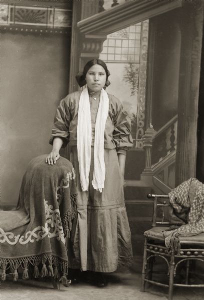 Studio portrait in front of a painted backdrop of a young Ho-Chunk woman posing standing with her right hand resting on a draped, overstuffed chair. She is wearing contemporary dress, including a hair bow and a long white silk scarf.