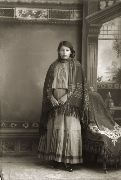 Studio portrait in front of a painted backdrop of a Ho-Chunk woman posing standing near a draped chair. She is wearing several rings, necklaces, a contemporary dark skirt and blouse, and a Racine Woolen Mills shawl over her shoulders.