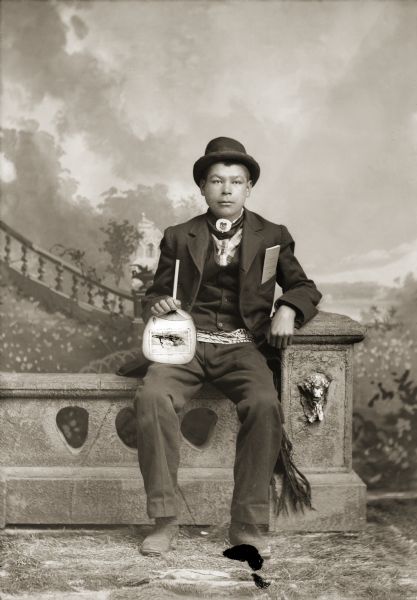 Studio portrait of Sam Wilson aka George Walkingcloud sitting on a stone wall. He is holding a paper fan in his right hand with an illustration of a boat. The fan appears to come from the 1892 Chicago World's Fair where a large number of Ho-Chunk performed. He also has a savings account book from a Black River Falls bank in his suit coat pocket, and is wearing a beaded belt, gorget, and hat. In the background is a painted backdrop.