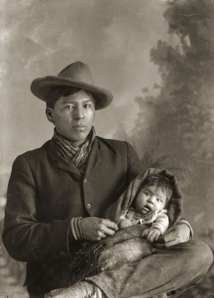Studio portrait of a Ho-Chunk man sitting with his leg crossed, and wearing a suit coat, bandana, and hat. He is holding his infant son who is wrapped in a blanket on his lap. Names, David Bow Littlesoldier (May Jhay Mau Nee Kah) and son, Simon Robert Littlesoldier (Wo No Ka Re Hunk Kah). In the background is a painted backdrop.