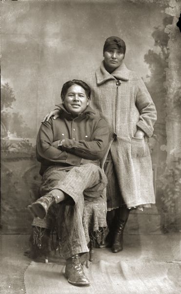 Studio portrait in front of a painted backdrop of a Ho-Chunk man, Louis Johnson (HaNuKaw), posing sitting on the left with his arms and legs crossed and wearing a wool shirt. A Ho-Chunk woman is standing next to him with her right hand on his shoulder. She is wearing a scarf wrapped around her head, a long coat, and boots.