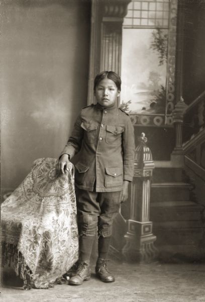 Studio full-length portrait in front of a painted backdrop of a Ho-Chunk boy, Esau Prescott, who is wearing a boarding school uniform. He is posing standing with his right hand on a chair draped with a fringed throw.