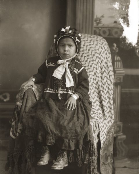 Studio portrait of a Ho-Chunk girl posing sitting on a cloth-covered chair in front of a painted backdrop. She is wearing several necklaces, file bracelets, a dark-colored dress, boots, and a bonnet tied with a light-colored ribbon. She is identified as Esther Dora White Yellowbank (HoChunkEWinKah), the daughter of Ulysses White (HoZheWaHeKah) and Queen Redwing-Winneshiek (Fannie Winneshiek) (WarConJarUKee).
