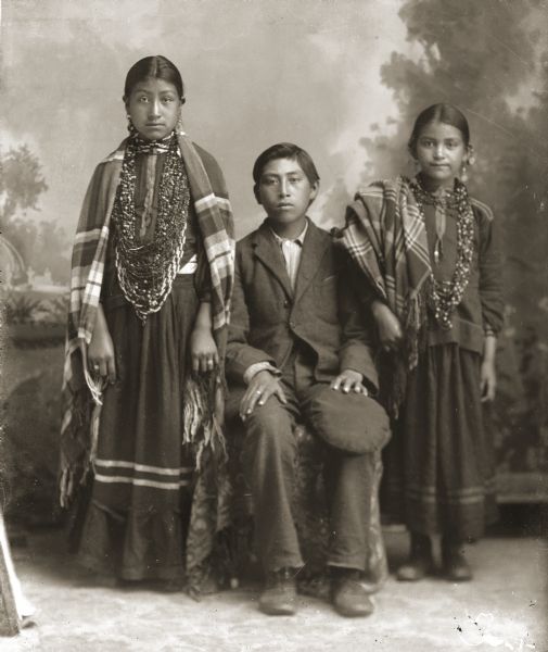 Studio portrait of three Ho-Chunk children posing in front of a painted backdrop. Identified as the children of Chief George Winneshiek (WaConChawNeeKah) and Rachel Funmaker Winneshiek (HockJawKooWinKah).  Clay Winneshiek (HunkMeNunkKah) is posing sitting holding a cap in his left hand on his left knee, and wearing a suit coat. Ellen Ella Winneshiek (TaChooHayWinKah) (left) and Fannie Winneshiek (HoChunkEHoNoNeekKah) (right) are posing standing on either side of the boy, and are wearing several necklaces and earrings with shawls over their shoulders.