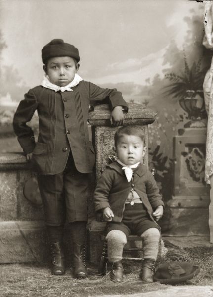 Studio portrait in front of a painted backdrop of a Ho-Chunk boy posing standing on the left with his left arm on a prop stone wall. He is wearing a winter hat, coat, and light-colored bandana. Another small Ho-Chunk boy is posing sitting on the right wearing a winter coat, short pants, wool leggings, and a light-colored bandana. A hat is on the floor by his feet.
