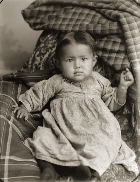 Studio portrait of a Ho-Chunk infant, John Stacy Jr. (HaGaKah), posing sitting on a pile of fabrics and wearing a dress. He is propped up on pillows to keep him steady for the camera. John Jr. was the son of John Stacy (ChoNeKayHunKah) and Martha Lyons-Lowe Stacy (KaRaChoWinKah).
