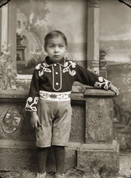Studio portrait of a small Ho-Chunk boy posing standing in front of a painted backdrop and a prop stone wall. He has his left arm resting on the wall, and is wearing a floral beaded shirt and beaded belt with short pants, leggings, and moccasins. Identified as Edwin (Ed Jr.) Greengrass (HoonkSepKah).