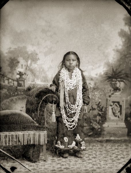Full-length studio portrait of a Ho-Chunk girl posing standing in front of a painted backdrop. She has her left hand resting on an overstuffed tasseled chair, and is wearing a rare floral beaded dress and many long strands of wampum.