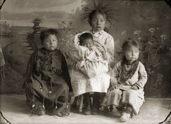 Full-length studio portrait in front of a painted backdrop of a Ho-Chunk boy posing sitting on the left wearing a fringed leather dress. In the center is a Ho-Chunk girl wearing a print dress and holding a Ho-Chunk infant in her lap. A Ho-Chunk girl is sitting on the right and is wearing several necklaces. Identified as the children of Frank Lewis (HaNaCheNaCooNeeKah)and Addie Littlesoldier Lewis Thunder (WauShinGaSaGah). From left to right: George Bryan Lewis (PatchDaHeKah), Amelia Emma Lewis (PatchKaRaWinKah) who is holding her brother Harry Ben Lewis (HooNaChaWeKah) and Mary (Pinkah) Lewis (KeSaWinKah).