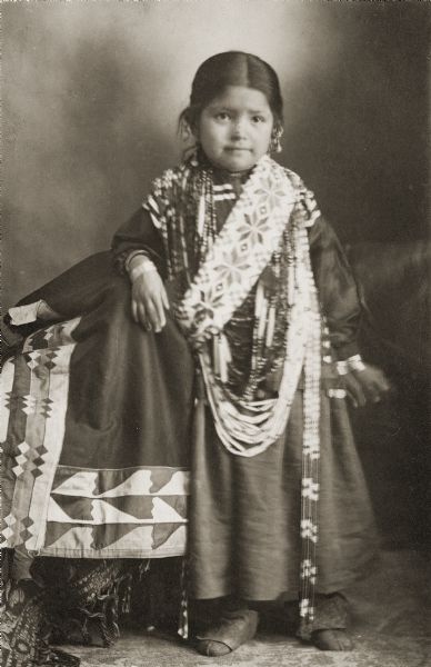 Full-length studio portrait of a young Mabel Mary Lonetree (ENooGah), the oldest daughter of Alec (Alex) Lonetree (NaENeeKeeKah) and Kate Winneshiek Lonetree (WauKonChawKooWinKah). She is posing with one arm resting on a covered chair and is wearing Ho-Chunk regalia including moccasins, earrings, and many necklaces and bracelets. Mabel Mary Lonetree was born on November 2nd, 1899 and died on March 30th, 1906 at the age of five from influenza. She was a younger sister to George Lonetree, the only child of Alec and Kate to survive the influenza epidemic. Their other siblings Howard and Anna Lonetree also died from the influenza epidemic; Anna first in January of 1905, Mabel Mary in March of 1906, and then Howard a month later.