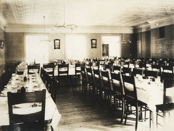 Dining hall at the Winnebago County Asylum. There are many chairs at tables set with tableware. Two large windows and a doorway are on the far wall, and light fixtures hang from a ceiling which has ceiling tiles.