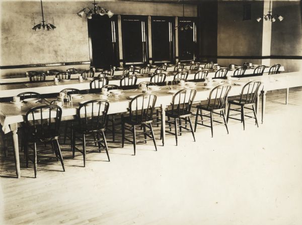Dining hall at the Winnebago County Asylum. In the foreground are spindle chairs pushed up against long tables set with tableware. Light fixtures are hanging from the ceiling, and on the back wall there is a doorway and three windows.