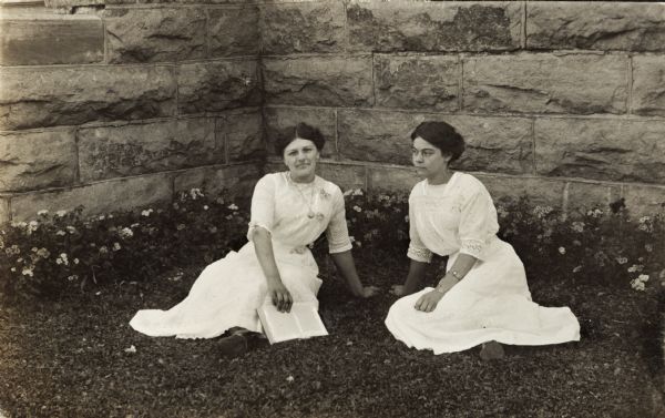 Two women sitting on the lawn of what appears to be the Winnebago County Asylum. Behind the women is a limestone block foundation lined with flowers. One of the woman is holding an open book in her lap.