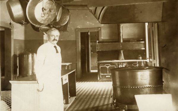 A man wearing an apron stands in a large kitchen located at the Winnebago County Asylum. Above him are pots and pans hanging from the ceiling. There is a large cauldron on the floor on the right, and behind it is an oven and stove.
