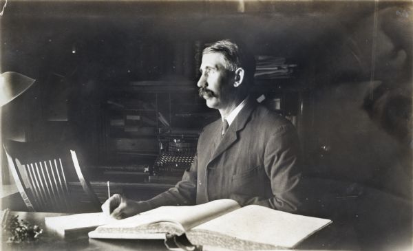 Portrait of E.E. Manuel, Superintendent of the Winnebago County Asylum. Manuel is seated at a desk with his head in profile while looking out the window. He has a ledger in front of him and behind him on a table is a typewriter.