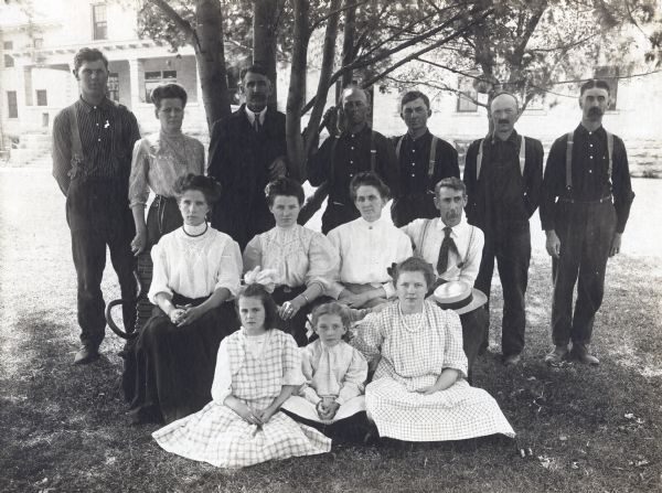 Outdoor group portrait of the Winnebago County Asylum administrative and nursing staff posed around a bench near a tree. Included in the portrait are the Superintendent, E.E. Manuel, his wife Olphene (Stromme) Manuel, Matron of the Asylum (both in the back row, far left side). Next to them is Superintendent, Elmer E. Manuel (third from the left, back row). In the front row, wearing a string of beads is Neita Faye (Manuel) Grueder, daughter of Elmer and Olphene and wife of George V. Grueder. Neita was Matron of the Asylum from 1929 to 1942. Visible behind the group is a building, most likely a Winnebago County Asylum building.