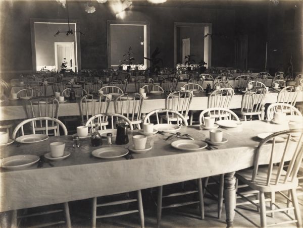 Dining hall at the Winnebago County Asylum. Spindle chairs are pushed up against long tables set with dishes. In the background are potted plants in front of three doorways leading into another room with more tables.