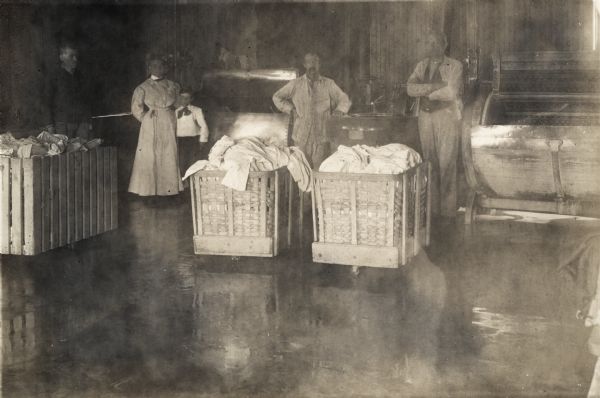 Staff members and one child posing among wheeled carts in the laundry facilities at the Winnebago County Asylum. In the background are two large laundry machines.