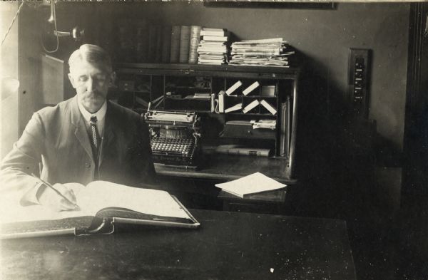 Portrait of E.E. Manuel, Superintendent of the Winnebago County Asylum sitting at his desk with a ledger in front of him. In the background is a typewriter, and what appears to be a telephone hanging on the wall.