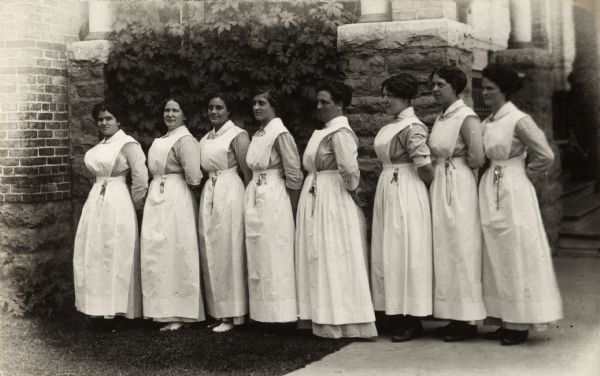Outdoor group portrait of the nursing staff at the Winnebago County Asylum. Eight nurses in uniform are lined up near the exterior of a brick building.