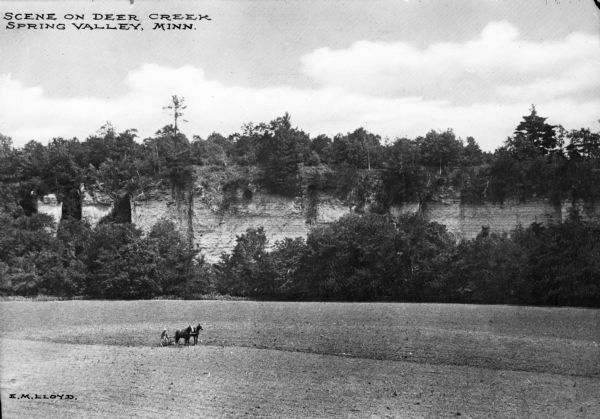 Elevated view of a farmer using a horse-drawn plow in a field near Deer Creek in Spring Valley, Minnesota. Tall rock formation can be seen on the edge of the field. Text at top left reads: "Scene On Deer Creek Spring Valley, Minn." and at bottom reads: "E.M. Lloyd."