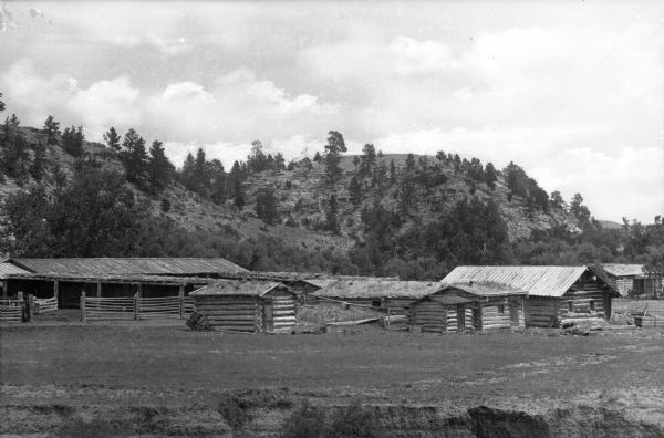 View of a ranch with log buildings and a fenced-in area. Hilly land covered with trees is in the background.
