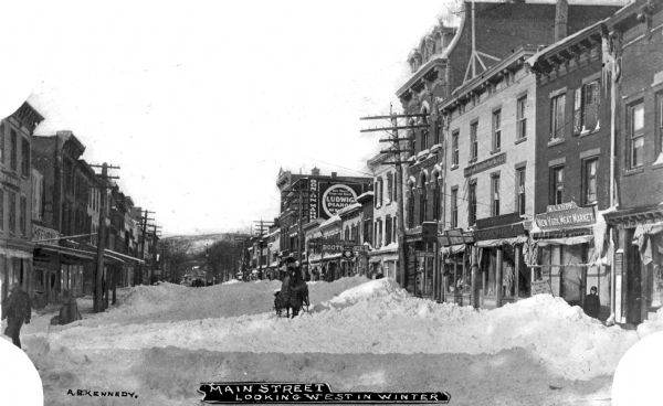 A view of Main Street after a snow storm. A horse-drawn vehicle can be seen making its way down the street and tall buildings line the snow-filled street. Text on photograph reads: "Main Street Looking West In Winter" and "A.B. Kennedy."