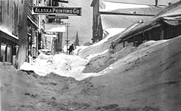 View of a city, filled with snow. A prominent business sign reads: "Alaska Printing Co: Photo Engraving." A pedestrian can be seen in the distance slogging through the snowdrifts that reach as high as the second story of buildings.