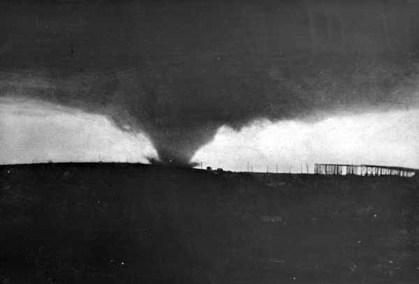 A tornado touching down in a field by a fence.  The funnel widens out dramatically on the mid-horizon into a dark, sky-filling cloud.