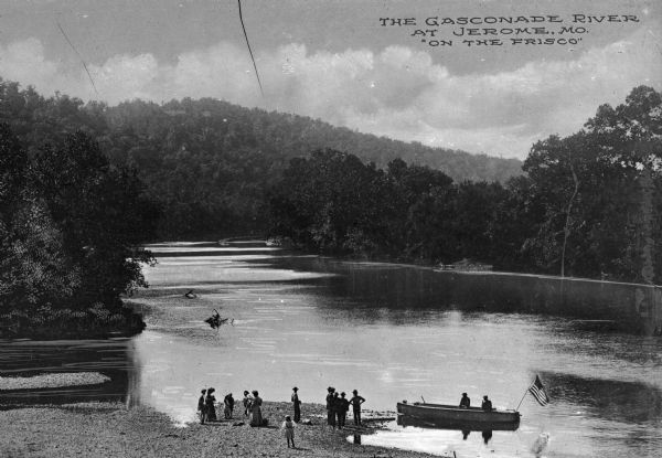 View of the Gasconade River and shore. A party of people are gathered near the water and a small rowboat with an American flag in it is pulled up alongside. Text on photograph reads: "The Gasconade River At Jerome, Mo. 'On The Frisco.'"