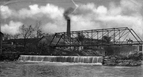 View of the steel Whittemore bridge passing over a dam. A man stands on the side of the dam and a horse and rider is crossing the bridge. On the shore stands an industrial building with a large smokestack.