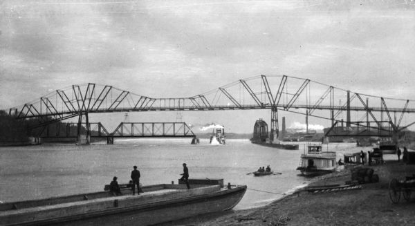 View of two bridges on the Mississippi River and the working river banks. A number of boats are in the water and cargo is stacked on the shore.