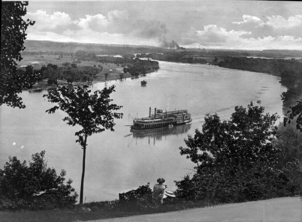 Elevated view of a steamboat on the Mississippi in the vicinity of Indian Mound Park. A woman sits on the riverbank in the foreground and the river winds through countryside into the distance.
