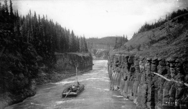 Elevated view of a flatboat traveling on the Yukon River through Miles Canyon in Yukon Territory, Canada. Growing on the land above the canyon are stands of pine trees.