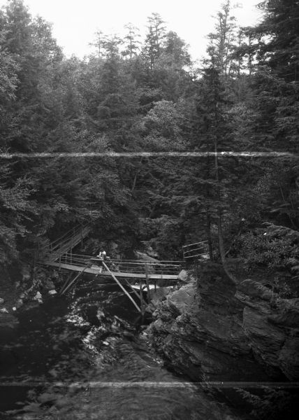 Elevated view of Winona Falls Gorge spanned by a small wooden bridge. Someone is resting in the middle of the bridge and a dense stand of pine trees surround the gorge.