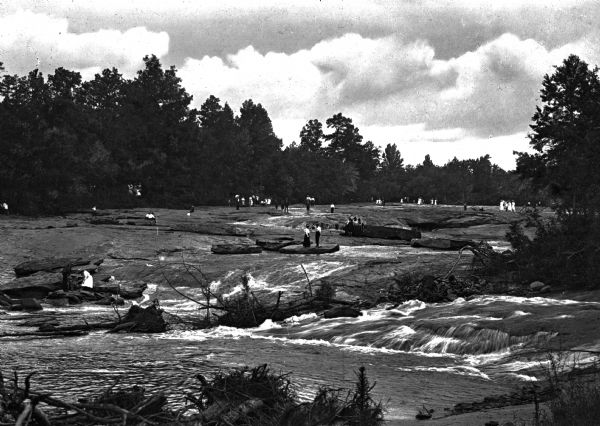 View toward people standing on large rocks in a river. The people are dispersed across the wide river and the river is lined by trees.
