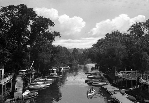 Elevated view of small boats tied up along a tree-lined stretch of the Blue River. Decks with stairs down to the boat slip areas stand on either side of the river.