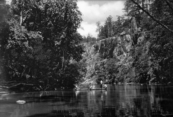 View of several people in a canoe on the little St. Frances River. A large rock formation can be seen in the background and trees grow along the riverbanks.