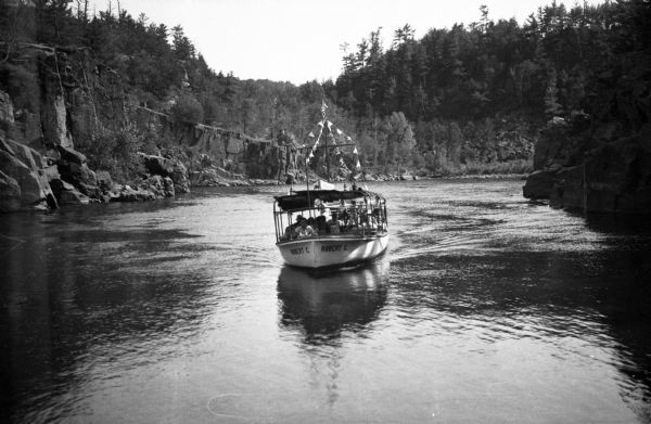 View toward the front of an excursion boat on the St. Croix River. The rocky shoreline is lined with pine trees.