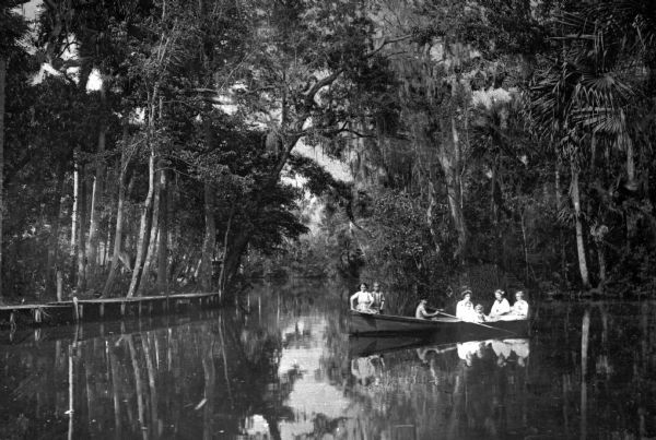 View of a group boating in the Everglades at the head of the Tomaka River. A wooden walkway is built along the treeline and Spanish moss hangs from branches above.